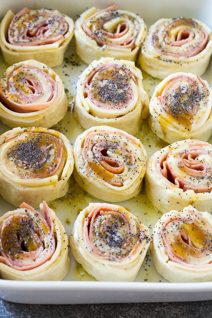 Ham and Cheese Pinwheels Recipe | Ham and Cheese Roll Ups | Ham and Cheese Rolls | Ham and Cheese Bread Rolls #ham #cheese #appetizer #pinwheels #ezlod Dough rolled up with ham and cheese and topped with a poppy seed butter in a ceramic dish.