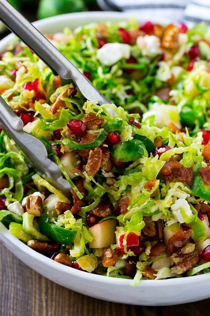 Brussels Sprout Salad Recipe | Shredded Brussels Sprouts Salad | Fall Salad #salad #brusselssprouts #bacon #thanksgiving #pomegranate #apple #dinner #ezlod A bowl of brussels sprout salad with tongs picking some up out of the bowl.