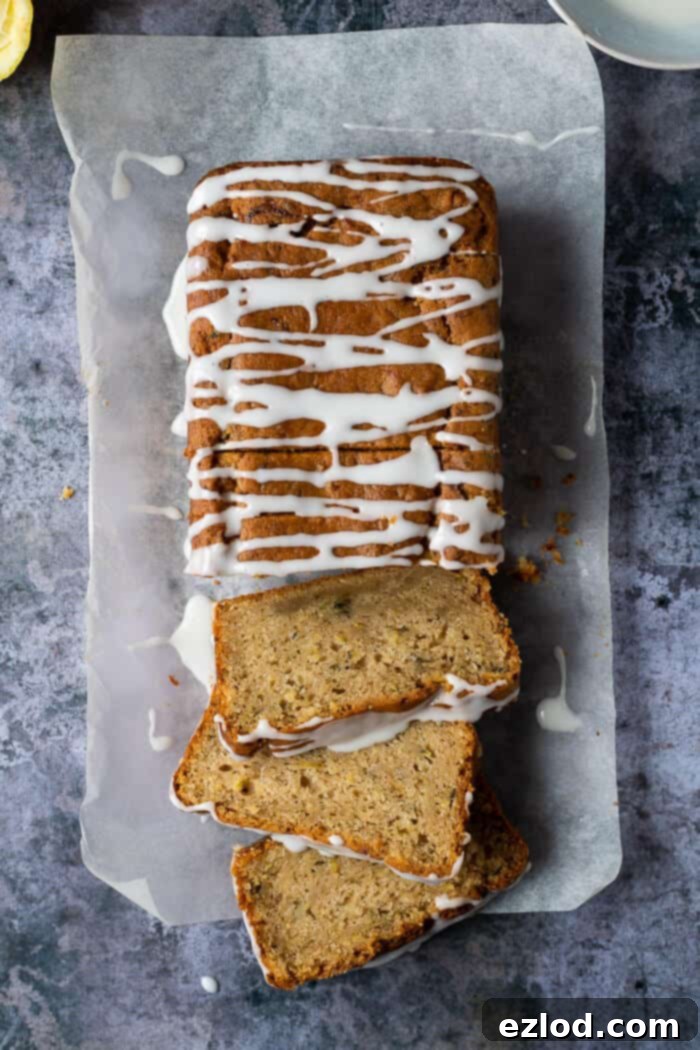 A whole, beautifully baked loaf of vegan courgette cake, delicately topped with lemon glaze, rests on baking parchment against a serene grey backdrop.