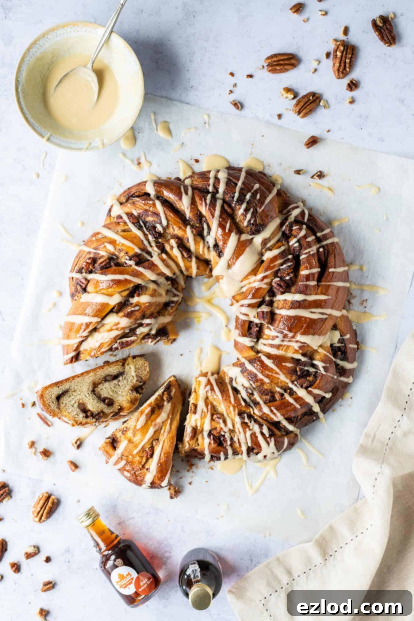 A beautifully baked vegan maple pecan bread wreath, golden brown and glistening with glaze, with two slices already cut out, resting on baking parchment. A small bowl of maple glaze, scattered pecan nuts, and mini bottles of maple syrup complete the inviting scene.
