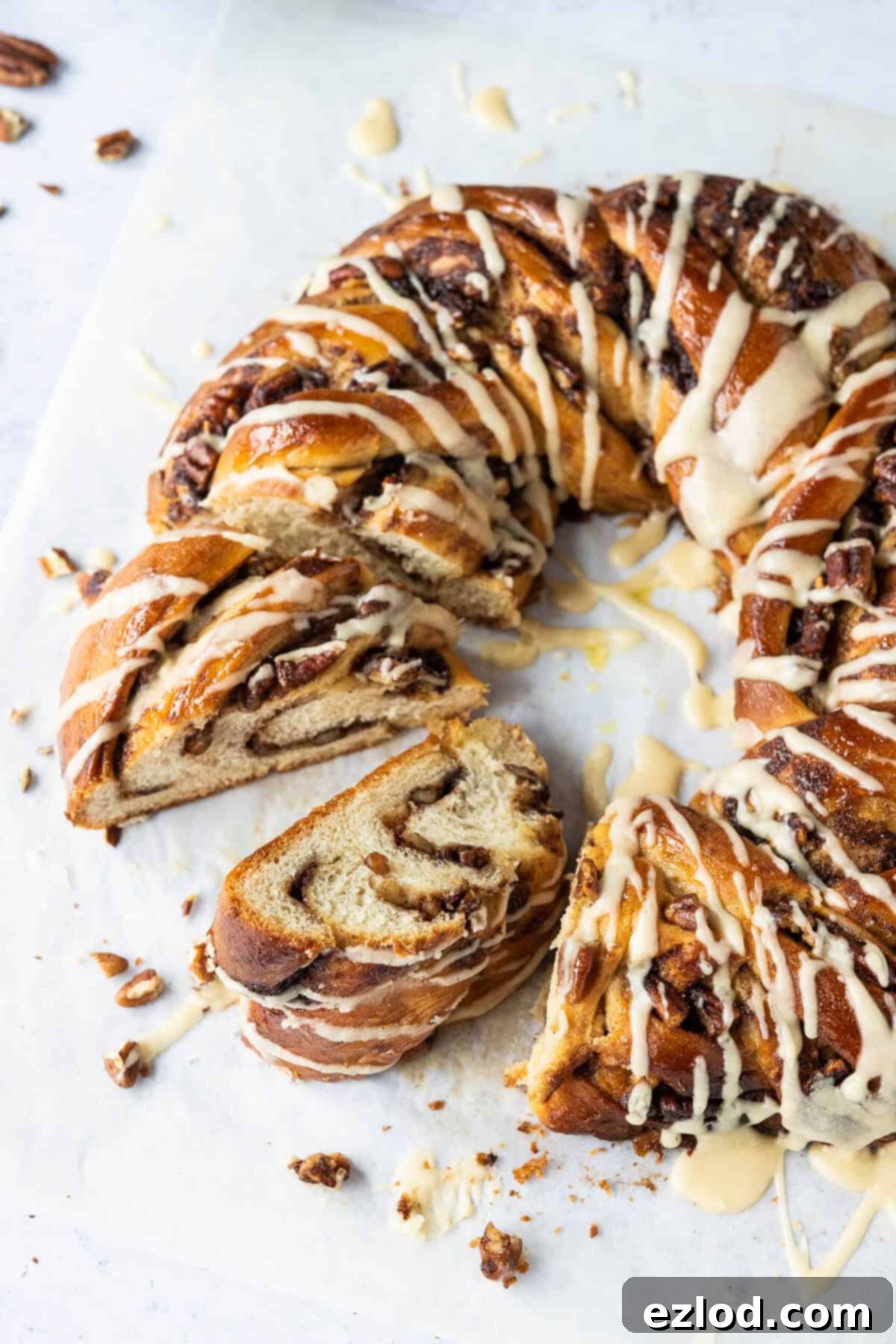 A close-up shot of a sliced vegan maple pecan bread wreath, revealing its swirl of cinnamon-pecan filling and generously drizzled with a rich maple glaze, all resting on a sheet of baking parchment.