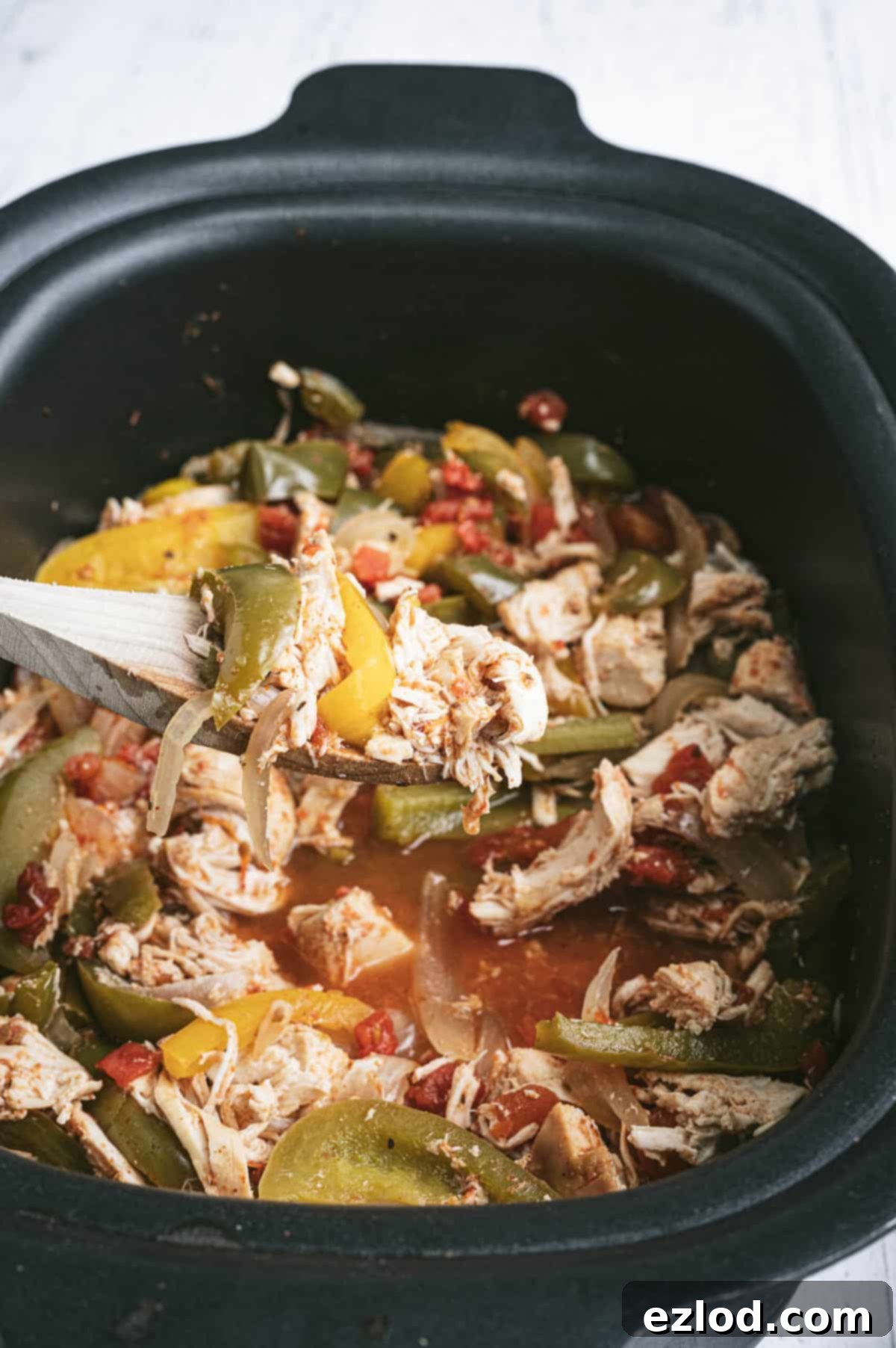 Effortless Slow Cooker Chicken Fajitas 4 A wooden spoon scooping out a portion of shredded Crockpot Chicken Fajitas with bell peppers and onions from a black slow cooker.