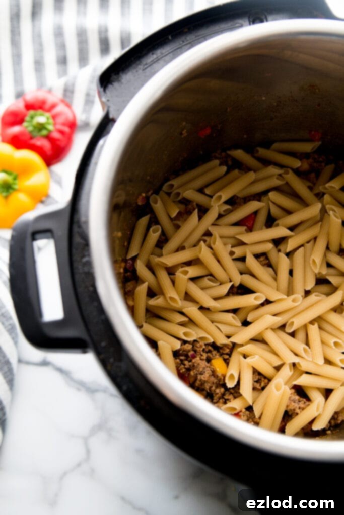 Overhead shot of gluten-free brown rice pasta, like penne, neatly layered over the seasoned ground beef and vegetable mixture in an Instant Pot, before liquids are added. This shows a crucial step for even cooking of the pasta in a one-pot meal.