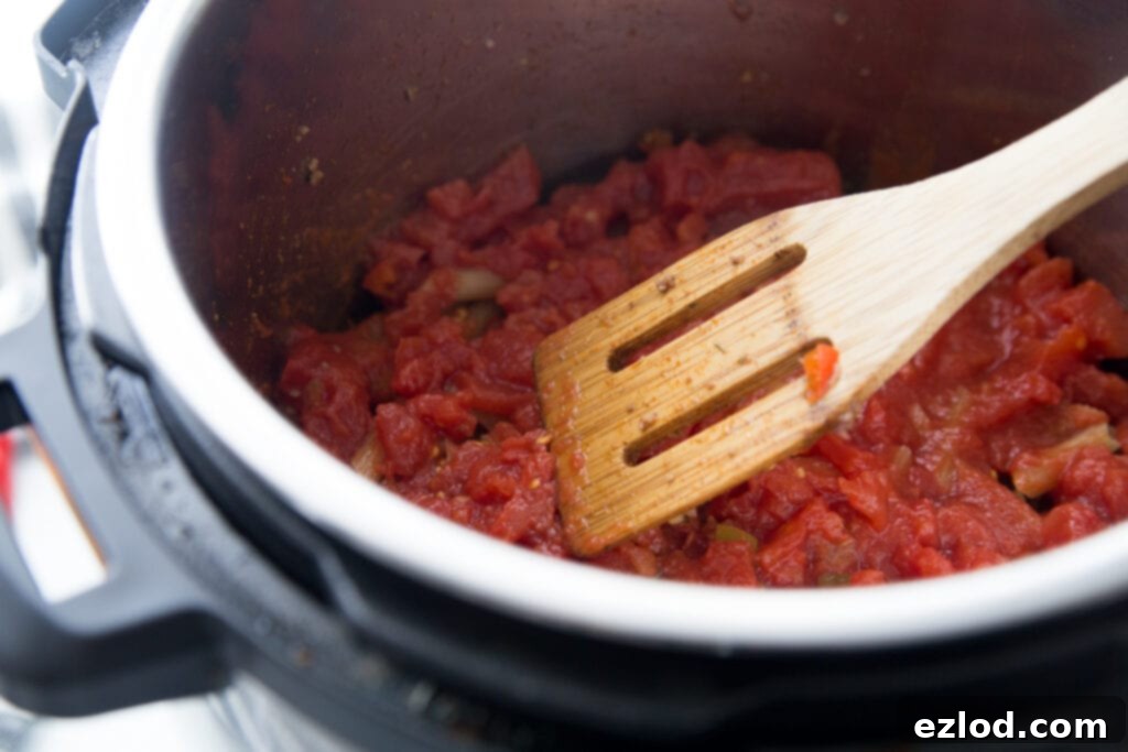 A close-up view of diced tomatoes with green chilies and rich tomato sauce being poured over layered pasta in an Instant Pot, ensuring even liquid distribution for a perfectly cooked Instant Pot Enchilada Pasta.