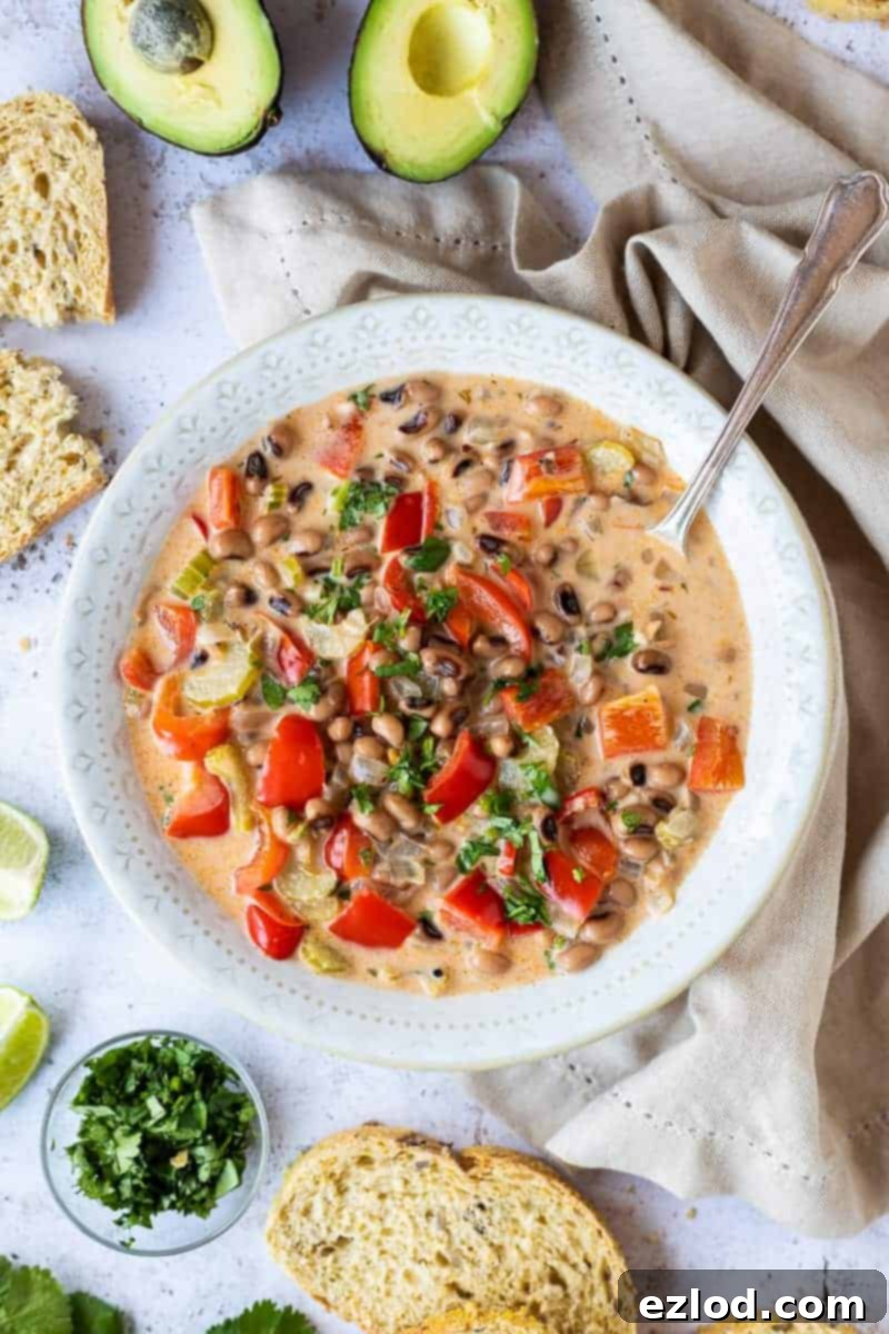 A white bowlful of vegan black-eyed bean soup surrounded by a cream coloured cloth, bread, avocado, lime wedges and chopped herbs. This quick and easy black eye bean, vegetable and coconut soup is a fast, delicious and healthy vegan option for lunch or dinner.