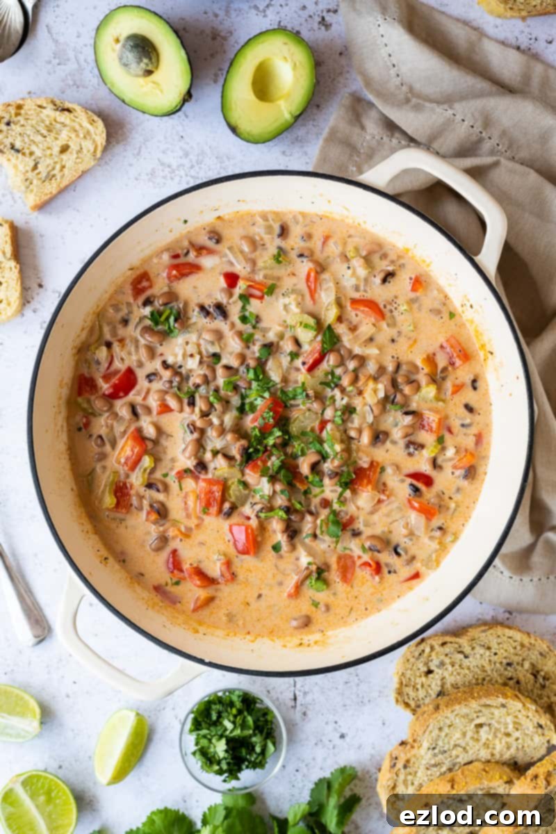 A white panful of easy black-eyed bean soup on a white background surrounded by sliced bread, avocado, a cream coloured cloth, lime wedges and chopped coriander. This quick and easy black eye bean, vegetable and coconut soup is a fast, delicious and healthy vegan option for lunch or dinner.