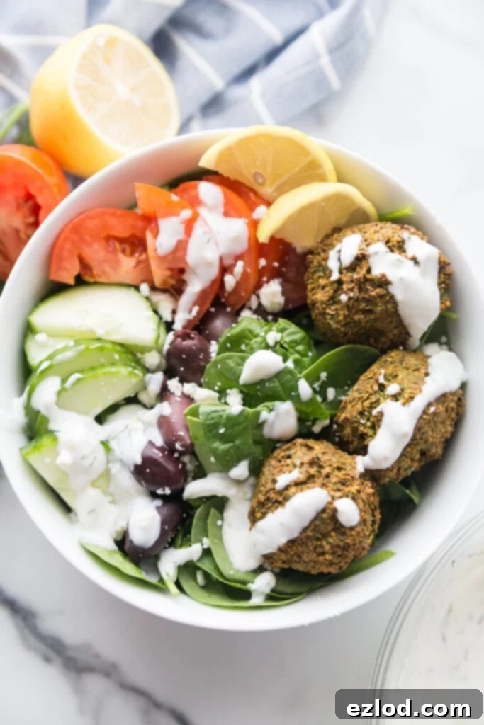 Overhead photo of large white bowl filled with Easy Falafel Bites and veggies including spinach, cucumbers, tomatoes, and olives, topped with tzatziki dressing with lemon wedges on the side.