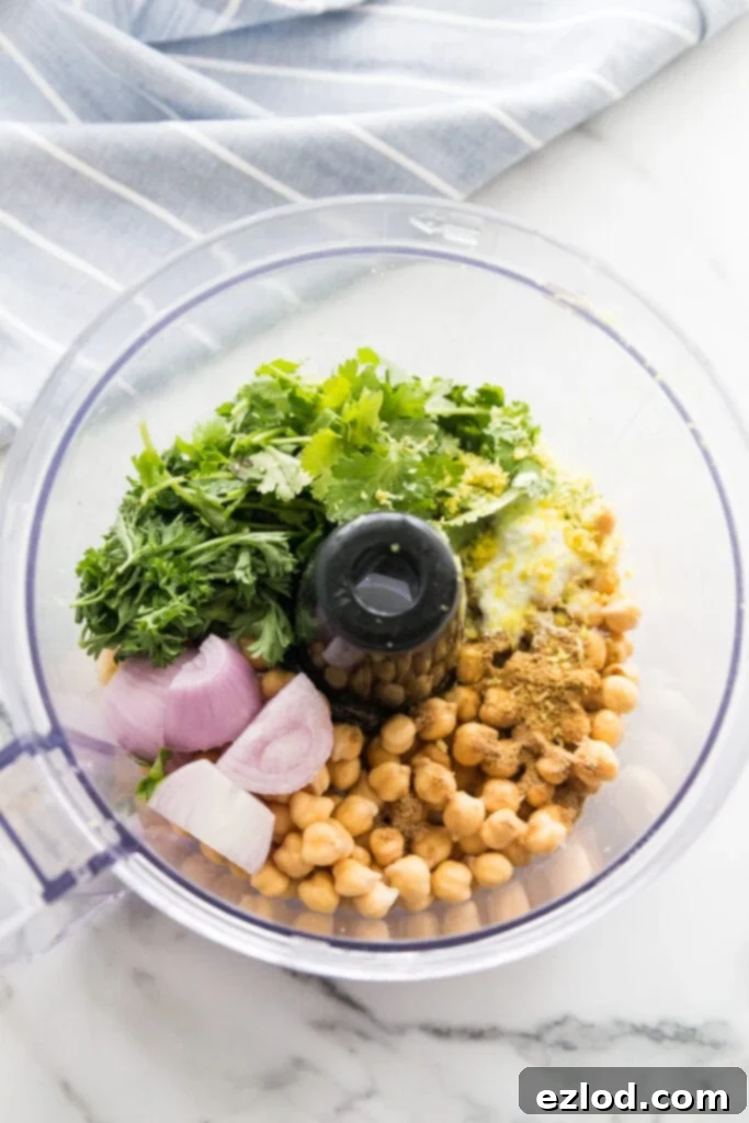 Overhead photo of food processor filled with ingredients for Easy Falafel Bites- chickpeas, shallots, cilantro, lemon zest, fresh parsley, and spices.