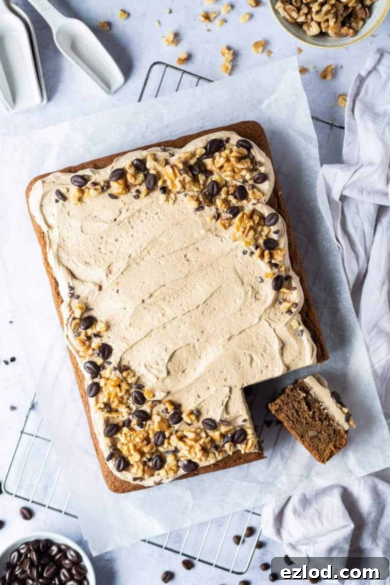 A delicious slice of vegan coffee and walnut traybake cake, perfectly cut from a larger sheet and displayed on baking parchment. The cake features a rich, moist crumb, topped with a generous swirl of light coffee buttercream, adorned with chocolate coffee beans and finely chopped walnuts. This crowd-pleasing plant-based dessert is resting on a wire rack, ready to be enjoyed.