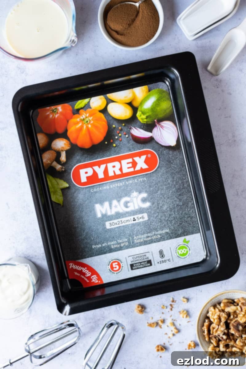 A high-quality Pyrex Magic Metal roasting tin sits centrally, surrounded by various baking ingredients and equipment. The scene includes flour, sugar, walnuts, coffee beans, measuring spoons, and a whisk, highlighting the preparation process for a delicious bake. The background suggests a kitchen setting, emphasizing the practical use of the bakeware.