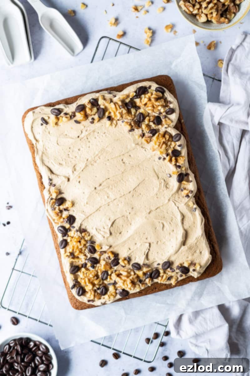 The beautifully iced and decorated vegan coffee and walnut cake, now a finished traybake, sits proudly on a wire rack against a soft grey background. It's garnished with an appealing array of chocolate coffee beans, crunchy walnuts, and delicate cacao nibs, highlighting its inviting and gourmet appearance.