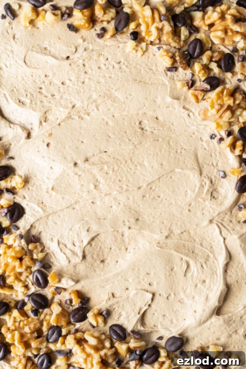 An inviting close-up of the luxurious coffee buttercream spread atop the vegan coffee and walnut traybake cake, artfully garnished with a mix of chocolate coffee beans, crunchy walnuts, and rich cacao nibs. This image highlights the cake's appealing texture and thoughtful decoration.
