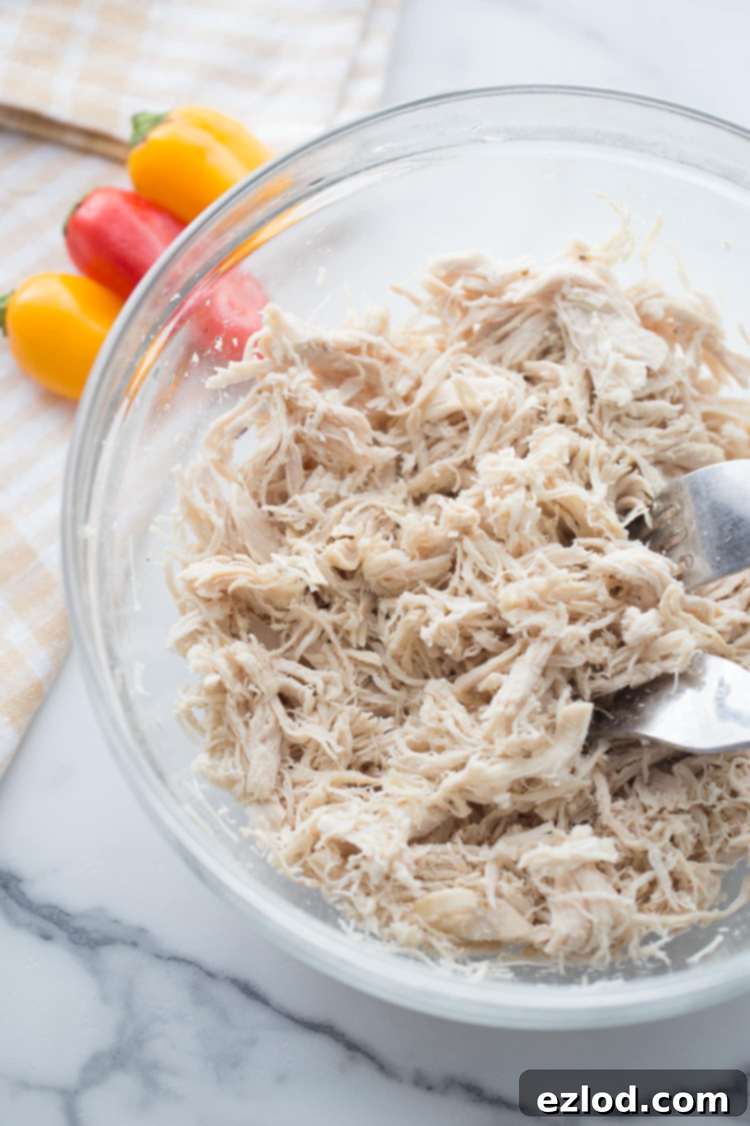 Fiery Buffalo Chicken Stuffed Peppers 5 Shredded chicken in a clear glass bowl with two forks, ready to be mixed, with colorful mini bell peppers blurred in the background.