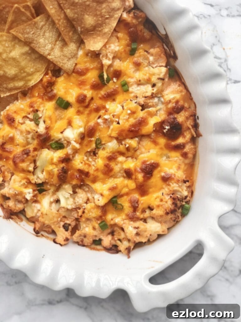 Overhead photo of golden-brown Buffalo Cauliflower Dip topped with crispy cheese in a white oval casserole dish, served with homemade tortilla chips, carrot sticks, and celery stalks on the side.