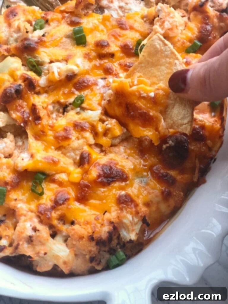 A woman's hand dipping a homemade tortilla chip into a hot, bubbly Buffalo Cauliflower Dip in a white casserole dish.