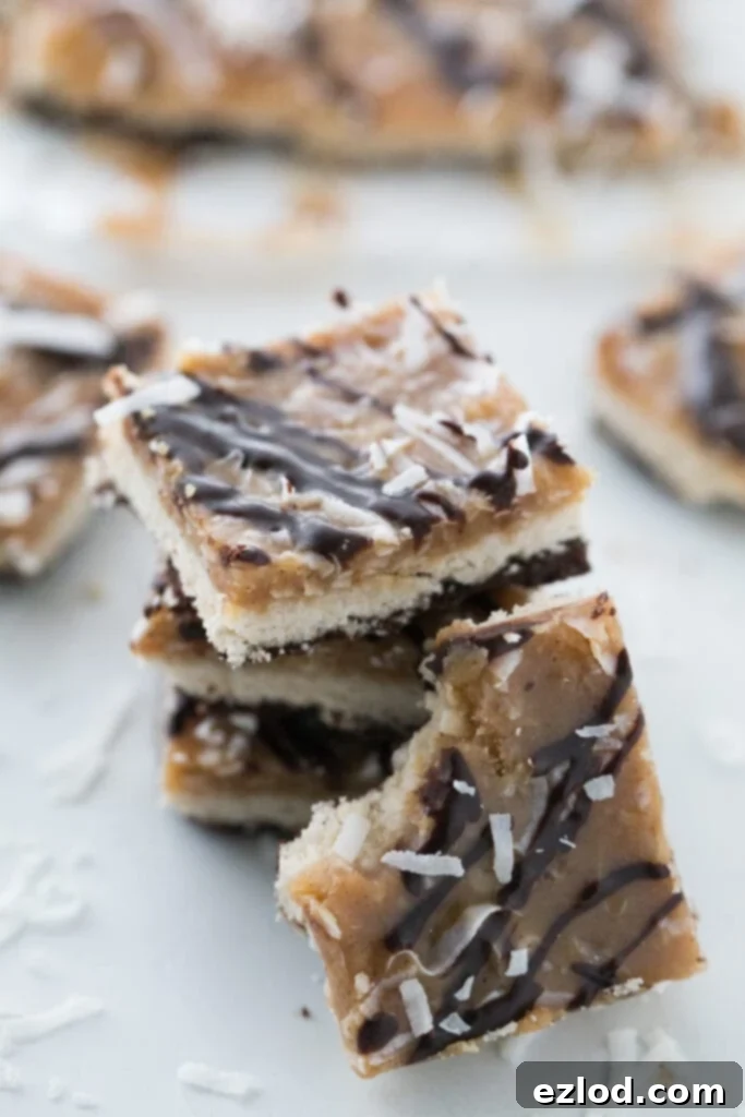 Close-up view of a stack of golden-brown Healthy Samoas Cookie Bars, featuring the distinct layers of coconut caramel and chocolate. Another bar leans against the stack, with more delicious treats softly out of focus in the background.