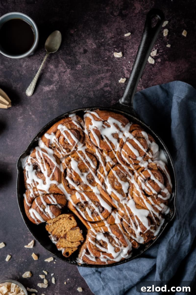 a cast iron skillet of vegan gingerbread cinnamon rolls, one sliced in half, on a dark background with candied ginger and a grey cloth.