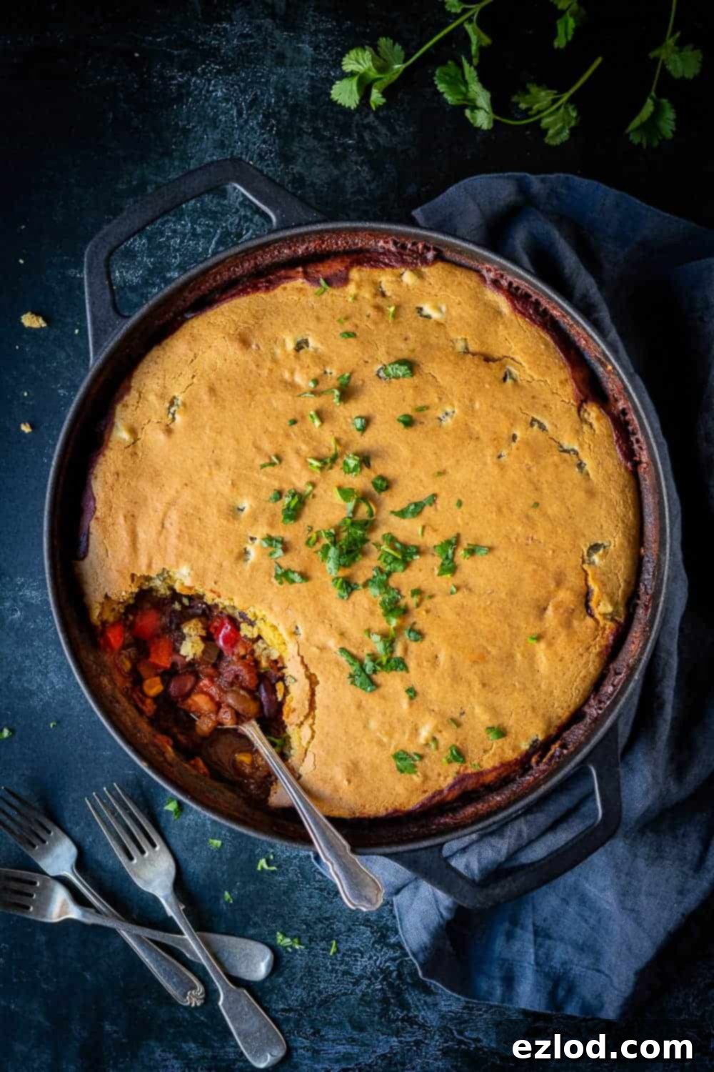 A beautifully cooked vegan chili cornbread pie presented in a black cast iron pan on a dark background, garnished with fresh chopped coriander. The golden cornbread topping looks invitingly fluffy.