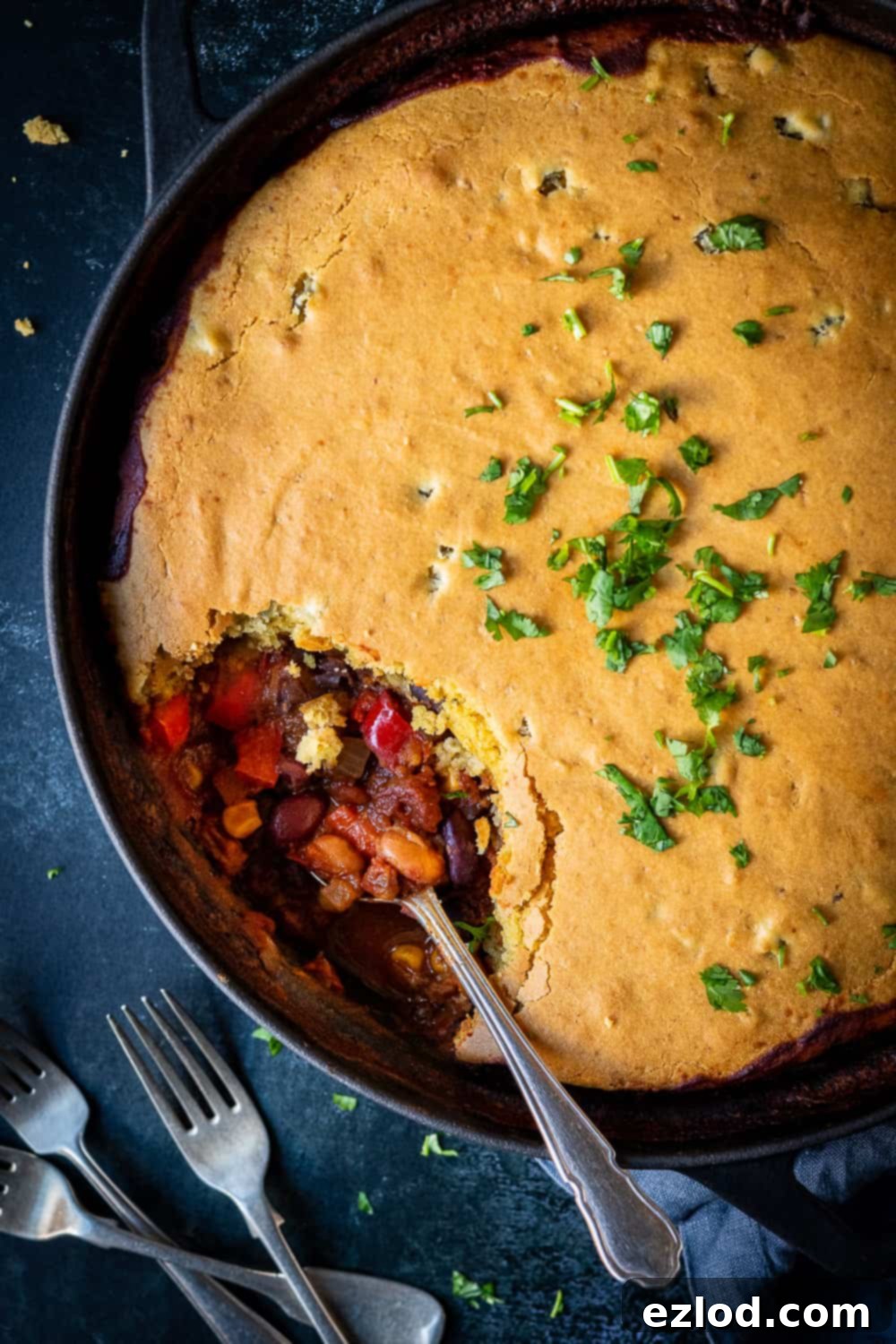 Vegan chili cornbread pie with a spoon in it in a black pan on a black background with a pile of forks, inviting to be served.
