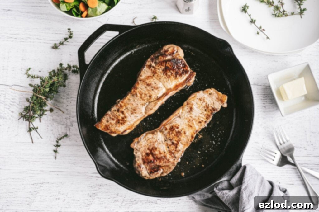 Overhead photo of New York Strip Steaks grilled in a cast iron skillet with thyme, butter, forks, and salad on the side.