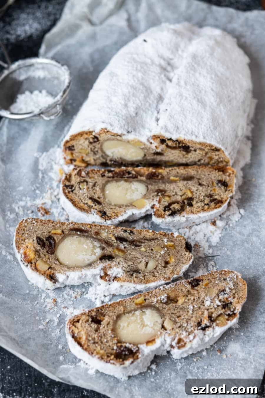 A whole loaf of vegan stollen on baking parchment, with a small sieve of powdered sugar in the background, ready for dusting.