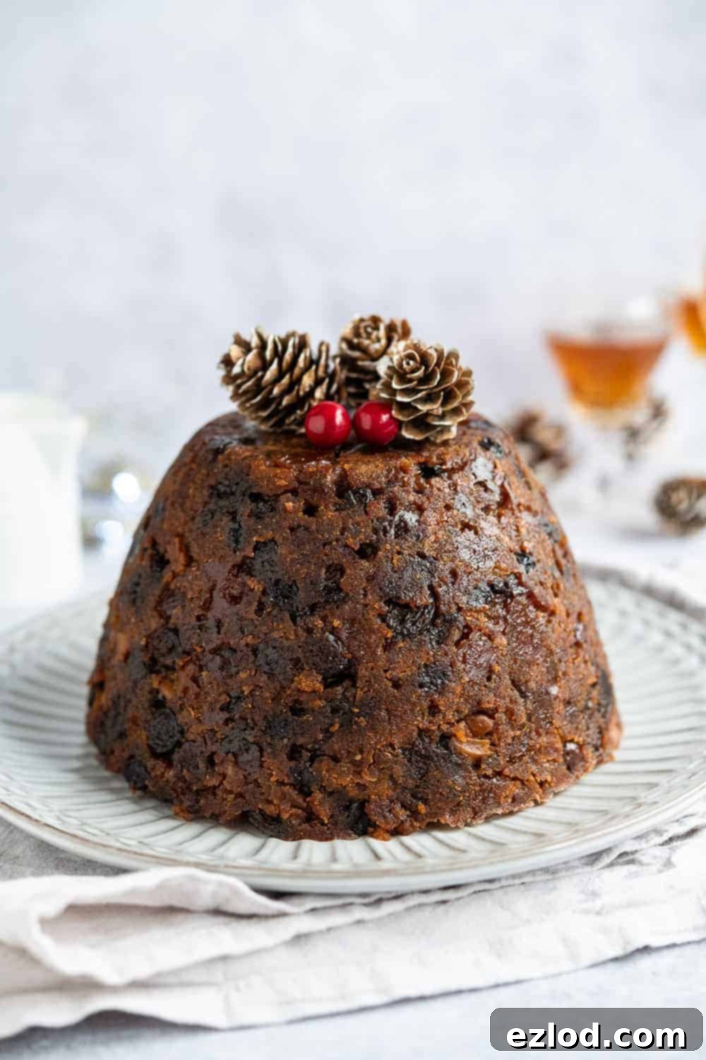 A beautifully presented vegan Christmas pudding on a white plate, set on a cream-colored cloth. The pudding is dark brown, adorned with festive pine cones and red berries, showcasing its rich, moist texture and inviting appeal. Ideal for a traditional vegan Christmas dessert.