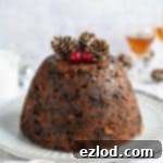 A close-up of a rustic vegan Christmas pudding on a white plate, resting on a light grey cloth, highlighting its rich texture and traditional appeal. Perfect for a festive dessert table.