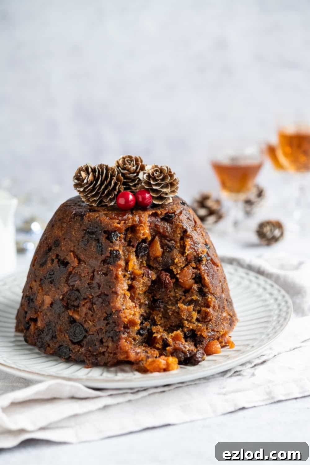 A slice of vegan Christmas pudding removed from the main pudding on a white plate, revealing its moist, fruit-filled interior. The background features blurred glasses of rum and decorative pine cones, emphasizing the festive, boozy theme of the dessert.