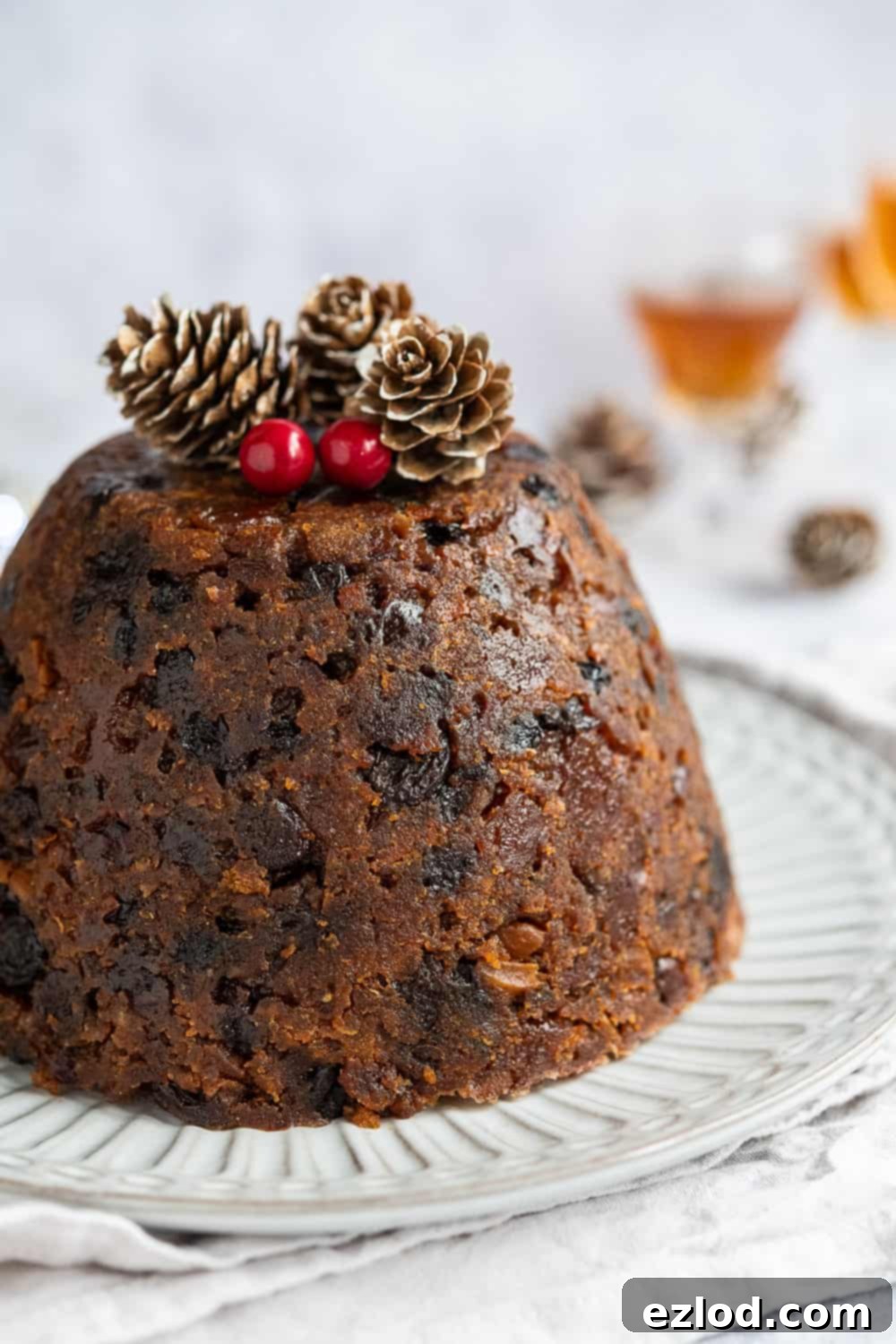 A close-up shot of a beautifully decorated vegan Christmas pudding, garnished with fresh pine cones and bright red berries, against a festive background. The image highlights the pudding's rich, dark color and its inviting, celebratory presentation.