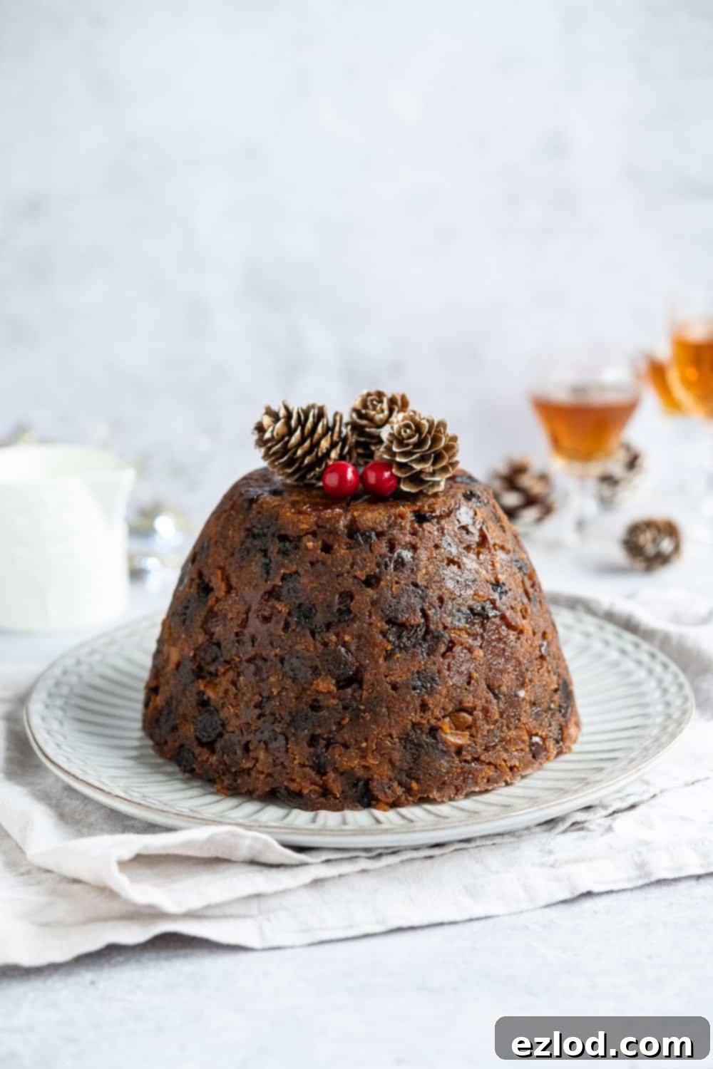 A beautifully arranged vegan Christmas pudding on a white plate, positioned on a grey cloth against a matching grey background. The scene is enhanced with festive elements like glasses of rum, a jug of creamy vegan custard, and decorative pine cones, illustrating perfect serving suggestions.