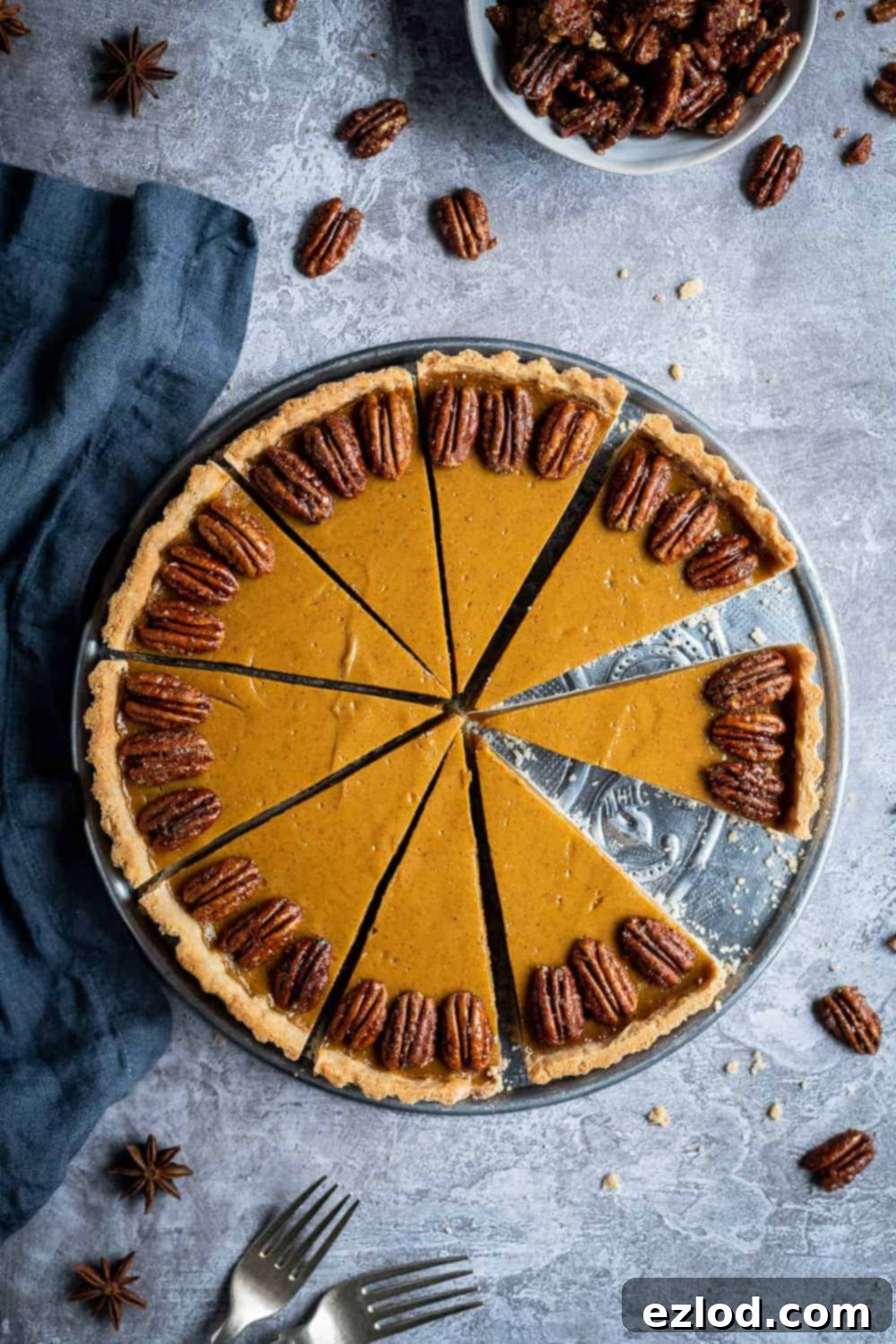 A beautifully baked vegan pumpkin pie, garnished with maple pecans, resting on a metal plate against a soft grey background. Perfect for Thanksgiving and autumnal celebrations.