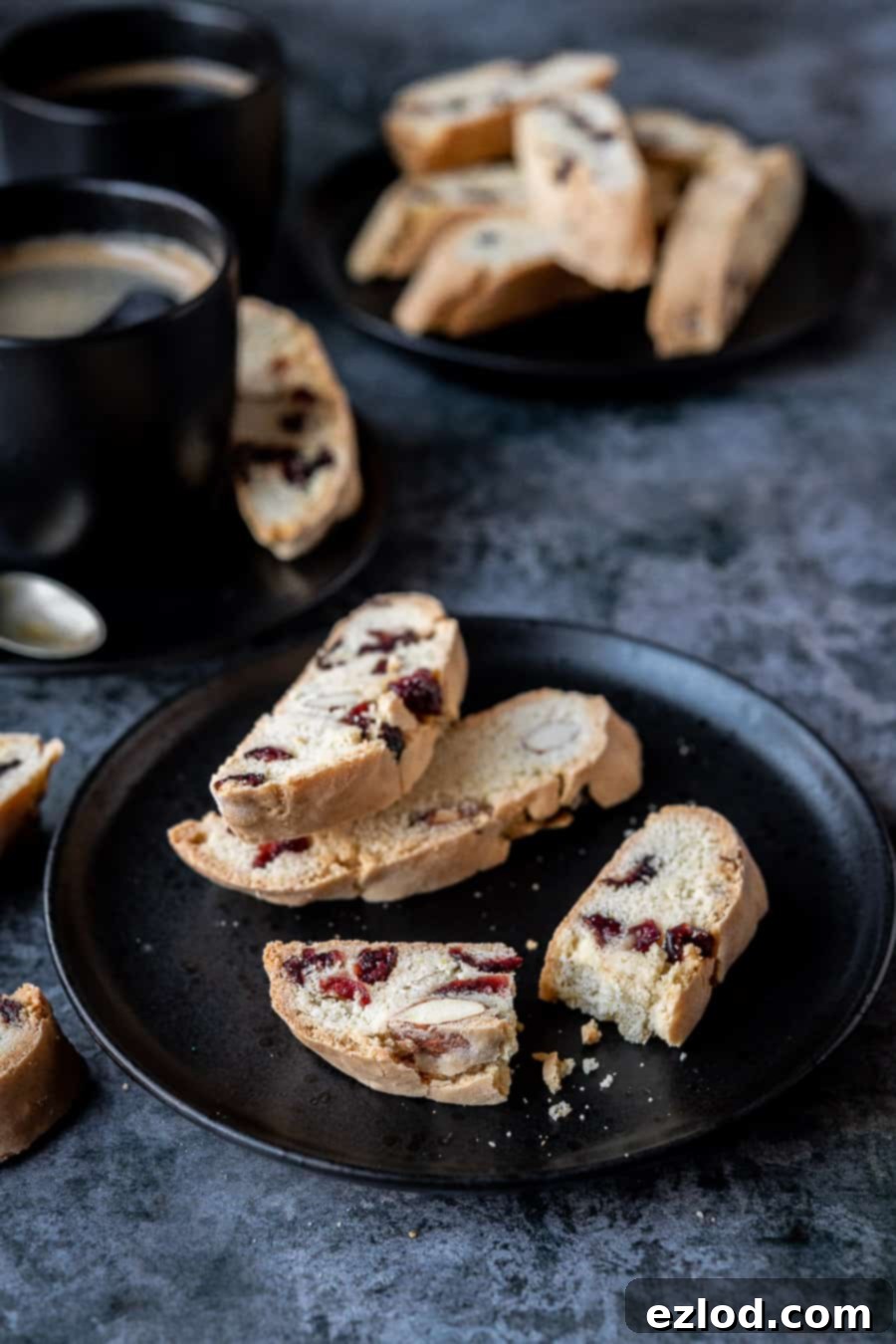Three vegan biscotti on a black plate, one broken in half, with a cup of coffee and another plate of biscotti in the background.