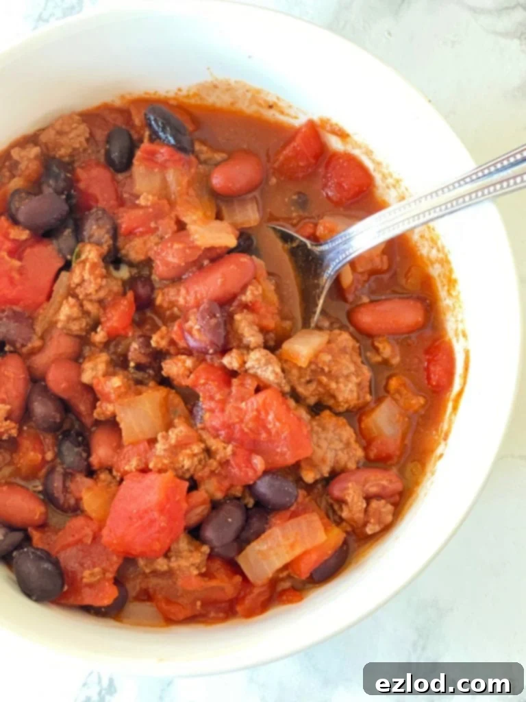 Close-up photo of a white bowl filled with delicious 5-ingredient chili, featuring canned tomatoes, black beans, kidney beans, ground meat, and onions. A spoon rests on the side.