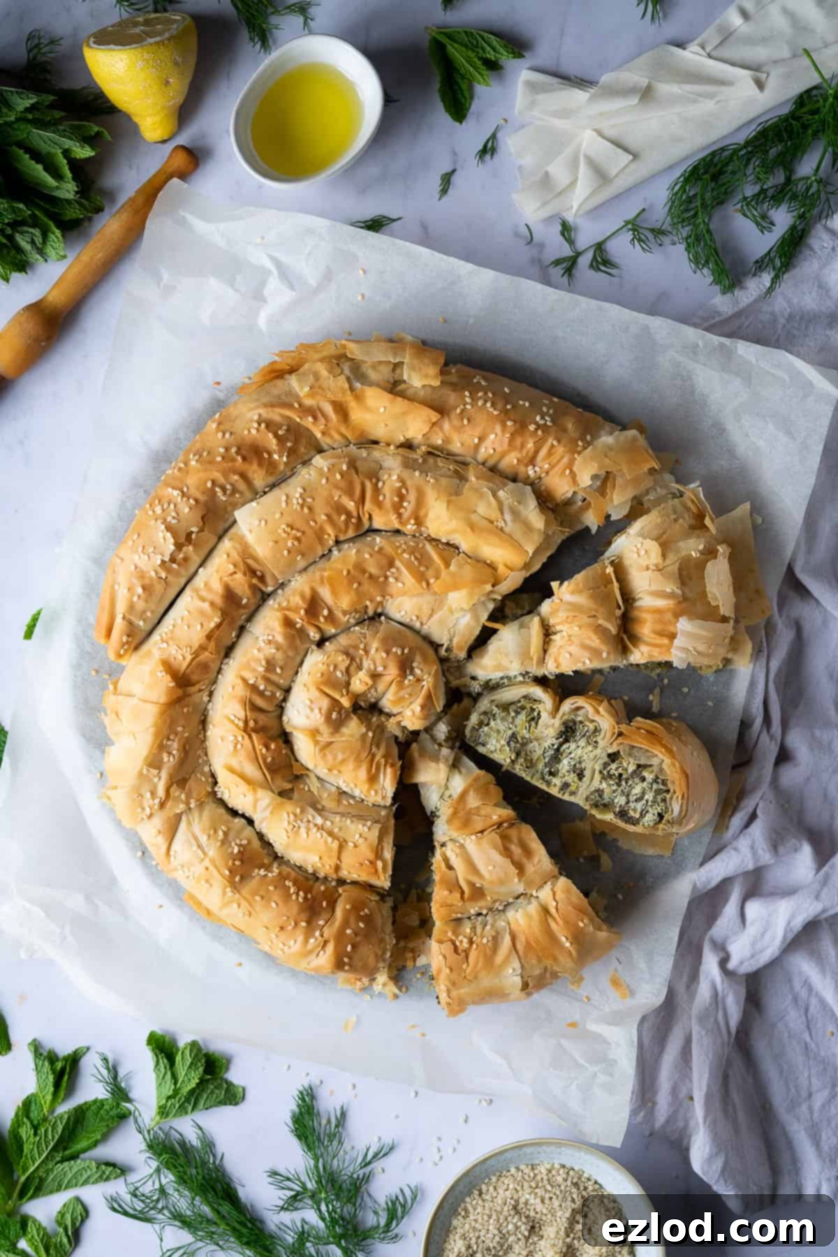 Sliced vegan spanakopita spiral on baking parchment, surrounded by fresh herbs and olive oil.