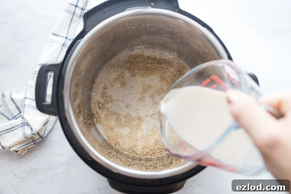 Overhead photo of uncooked rice in an instant pot with milk being poured into the pot from a clear measuring cup. On the side is a blue and white plaid kitchen towel on a white marble surface.