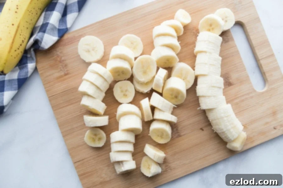 Sliced bananas on a wooden cutting board on a white marble surface. In the corner you can see part of a banana on a blue and white gingham towel.