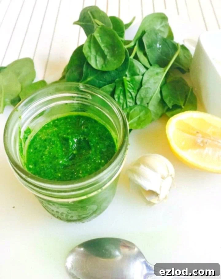 Side view of a mason jar full of homemade spinach pesto sauce on a white cutting board with a white wooden background too. Next to the jar are some ingredients to make it- a pile of spinach leaves, half of a lemon, half of a bulb of garlic, and an overturned spoon. 