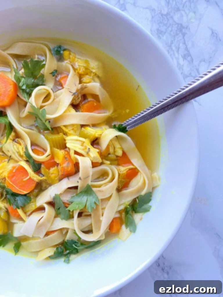 Overhead view of a bowl of Chicken Noodle Soup with Gluten free egg noodles, chicken, sliced carrots, diced celery, cilantro garnish, and beautiful yellow broth. There is a spoon in the white bowl on a white marble background.