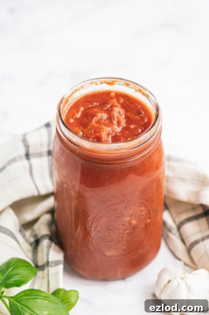 High angle of a large mason jar full to the brim of homemade tomato sauce. Near the jar is a white and blue plaid towel, some fresh basil, and a bulb of garlic on a white surface.