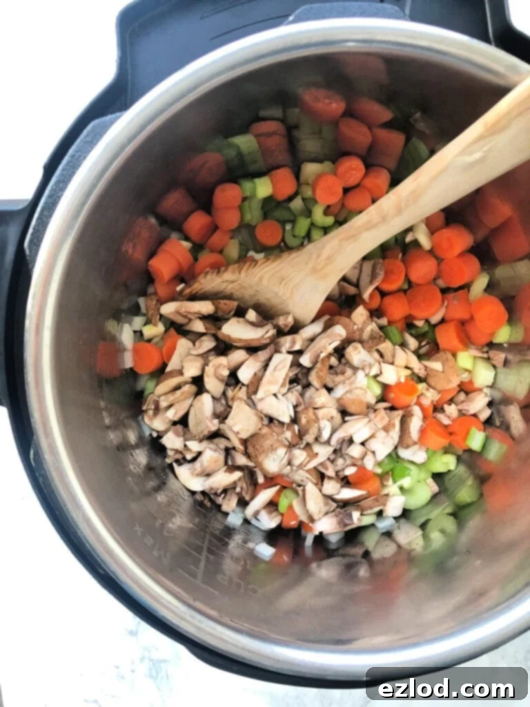 Overhead view of an instant pot full of chopped veggies- carrots, celery, onion, mushrooms- with a wooden spoon, ready to stir.