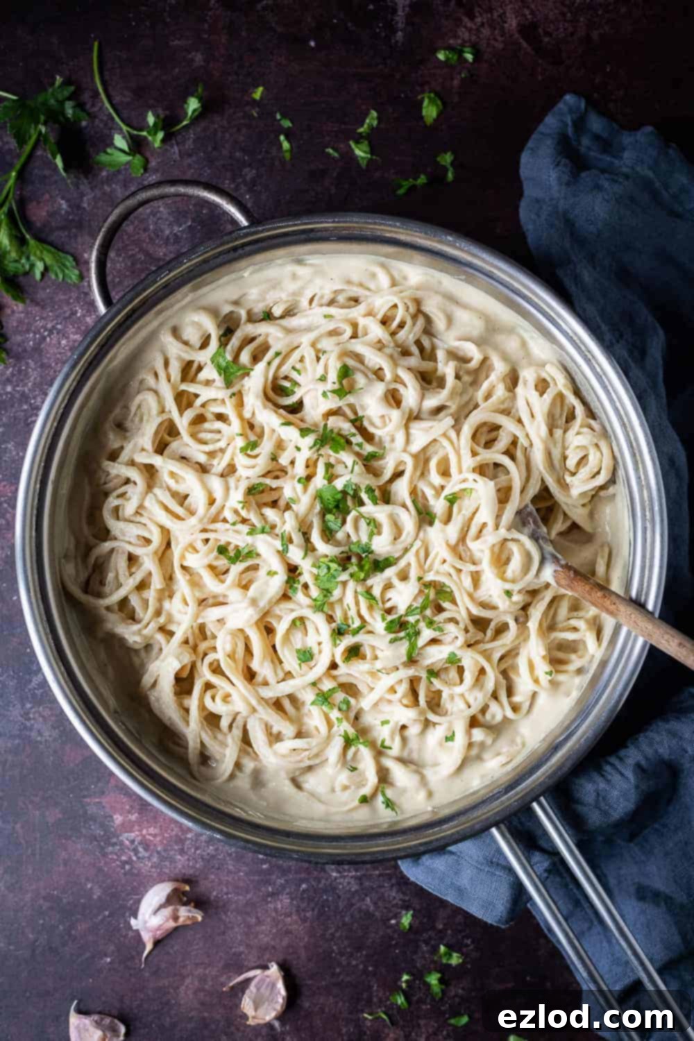 A pan filled with luscious, creamy vegan cauliflower alfredo pasta, garnished with fresh parsley, on a dark brown background. Garlic skins and a grey cloth add to the rustic appeal.