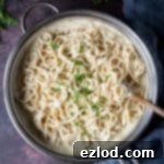 A panful of creamy vegan cauliflower Alfredo pasta on a dark brown background with parsley, a grey cloth and garlic skins, illustrating the finished dish.