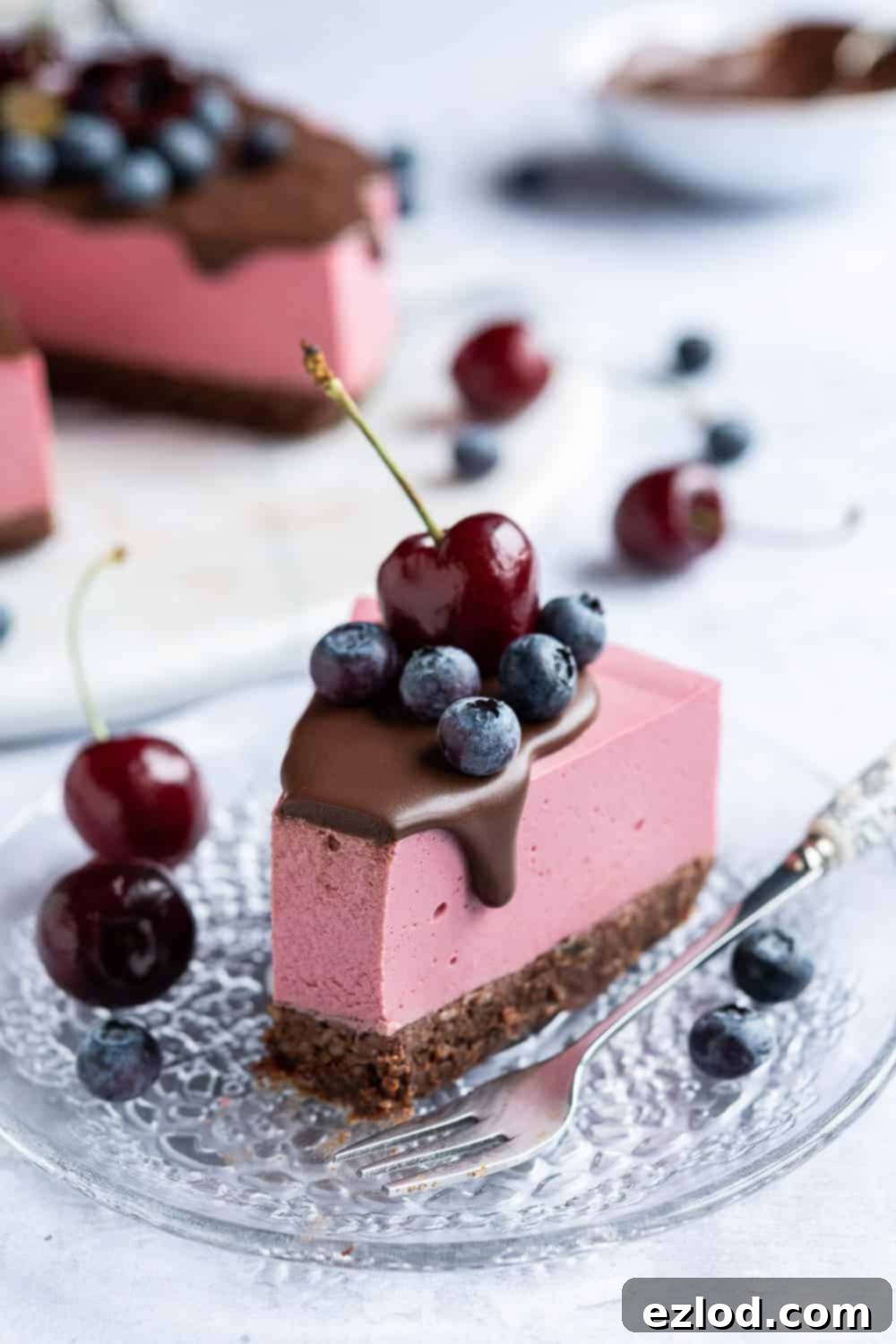 A slice of vibrant no-bake vegan chocolate cherry cheesecake topped with fresh blueberries and a cherry, served on a elegant glass plate with a forkful removed, showcasing its creamy texture.