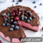 A slice of no-bake vegan chocolate cherry cheesecake on a glass plate with a fork, decorated with fresh blueberries and a cherry, alongside the full cheesecake topped with berries.