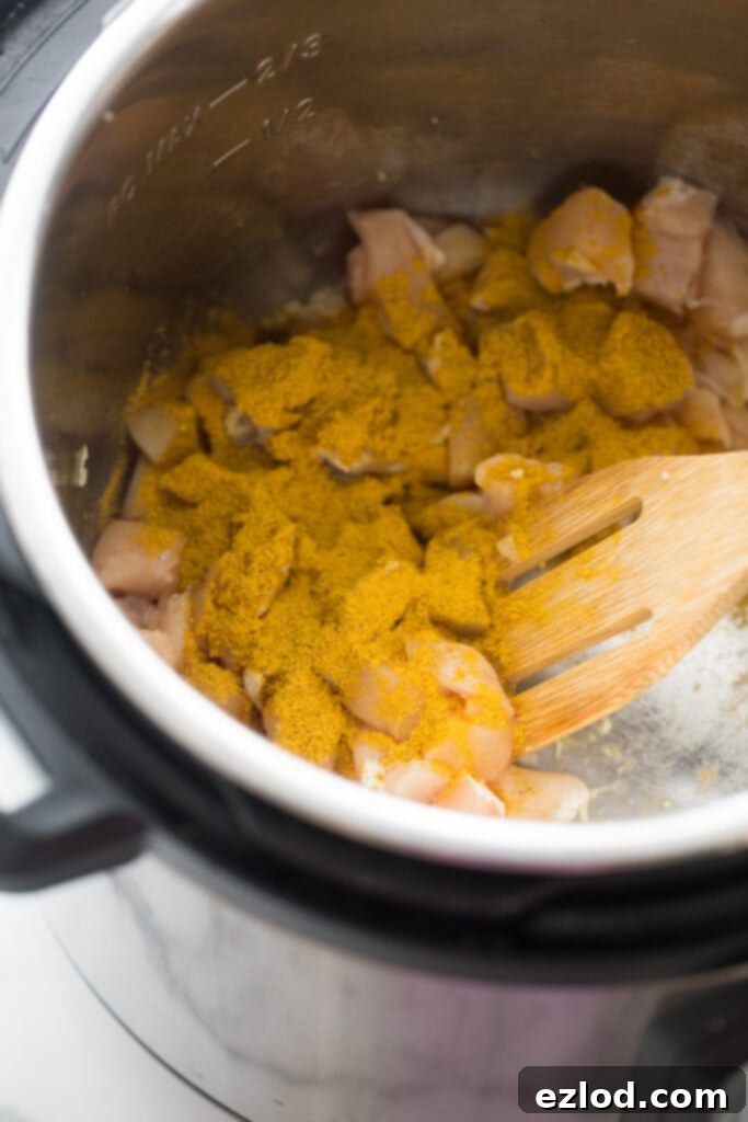 An overhead shot showing diced raw chicken pieces, thoroughly coated in vibrant yellow curry seasoning, resting in the Instant Pot, with a wooden slotted utensil ready for stirring.