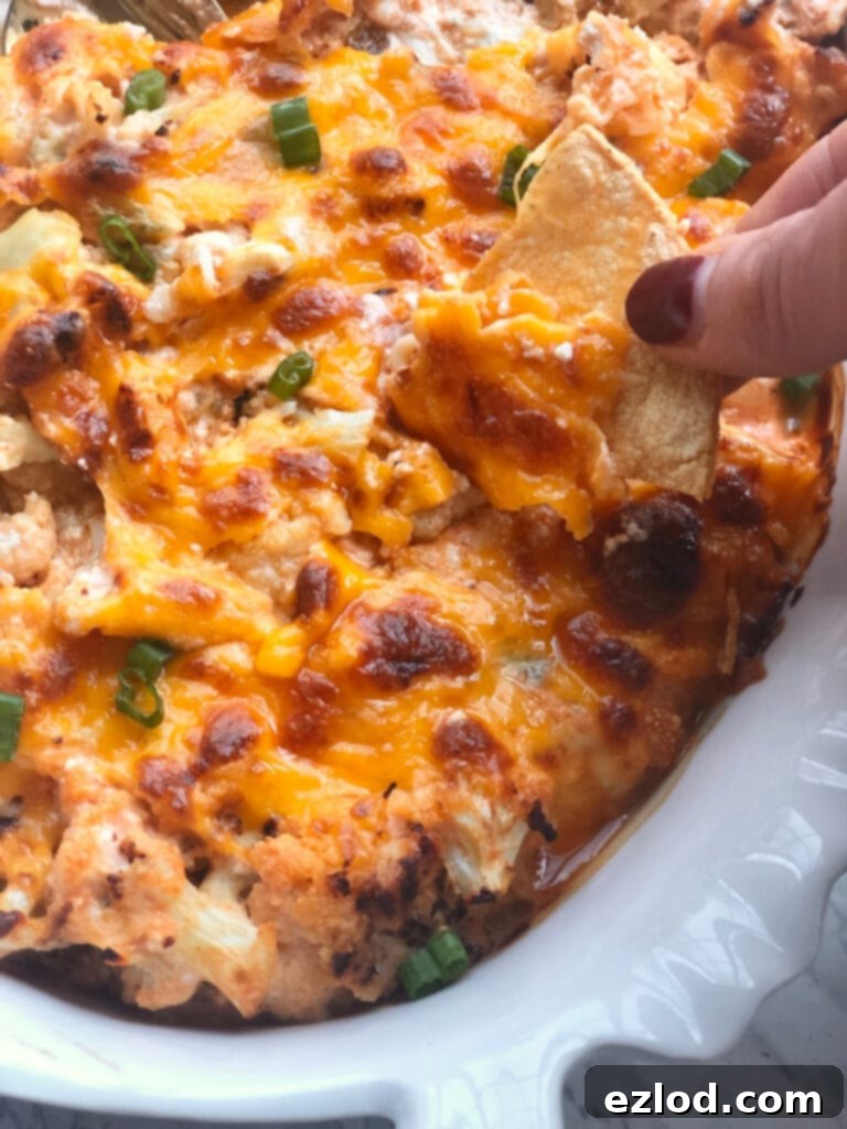 A woman's hand with painted fingernails is dipping a homemade tortilla chip into buffalo cauliflower dip. The dip is in a white oval serving dish with crispy, bubbly cheese on top and garnished with green onions- a great savory snack option.