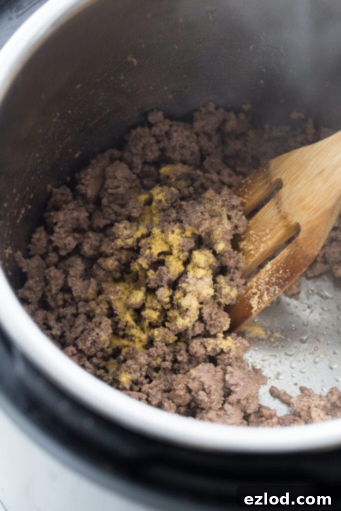 Overhead view of an Instant Pot full of browned ground beef, topped with spices. There is a slotted wooden spoon stirring the meat.
