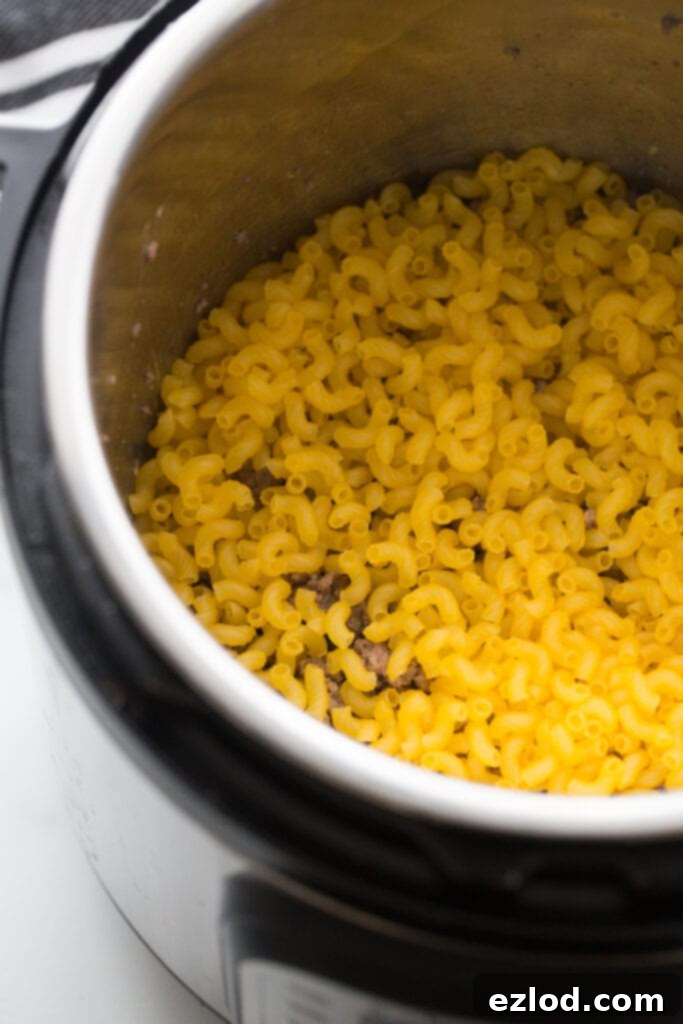 Overhead view of an instant pot full of a layer of raw gluten free elbow noodles on top of cooked hamburger meat, in process of making a healthy hamburger helper recipe.