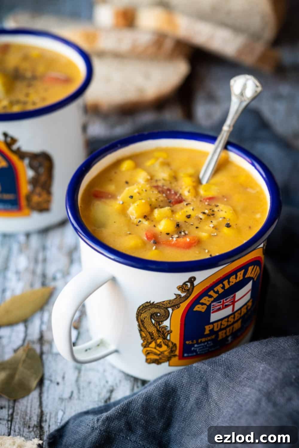 vegan corn chowder in a blue and white enamel mug with a silver spoon and sliced bread and a grey cloth in the background.