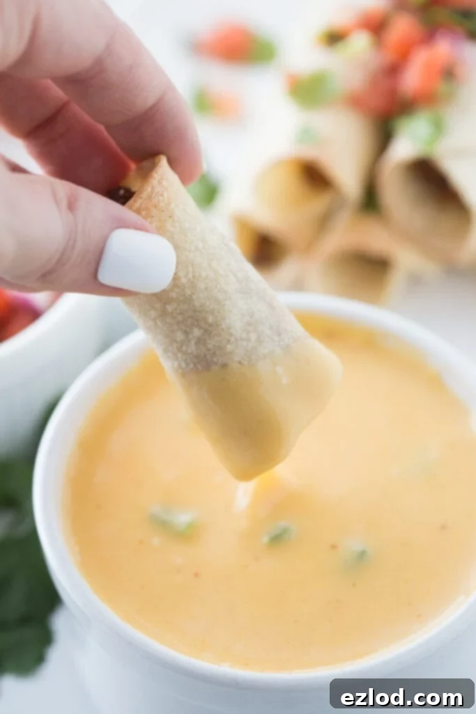 Woman's hand with white nail polish is holding a beef taquito and dipping it into a bowl of cheese sauce. In the background, out of focus, is a stack of taquitos topped with homemade salsa.